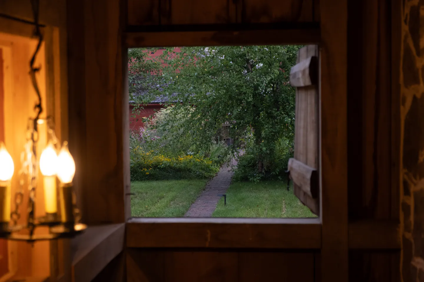 View through a barn window at dusk: candle lamps inside, a brick path and red barn beyond